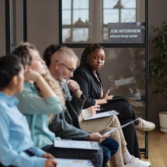 African young woman reading a contract while sitting with other people in hallway at office and waiting for her turn to job interview